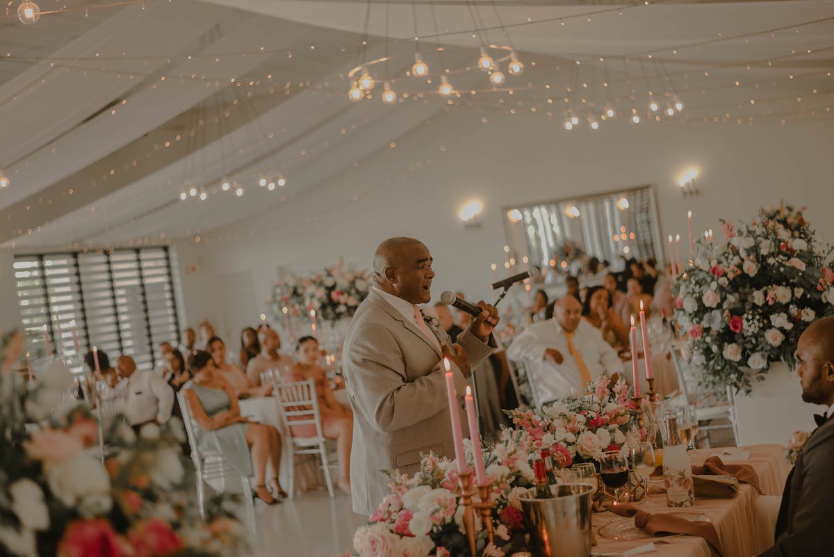 A man gives a speech at a wedding reception surrounded by guests and floral arrangements
