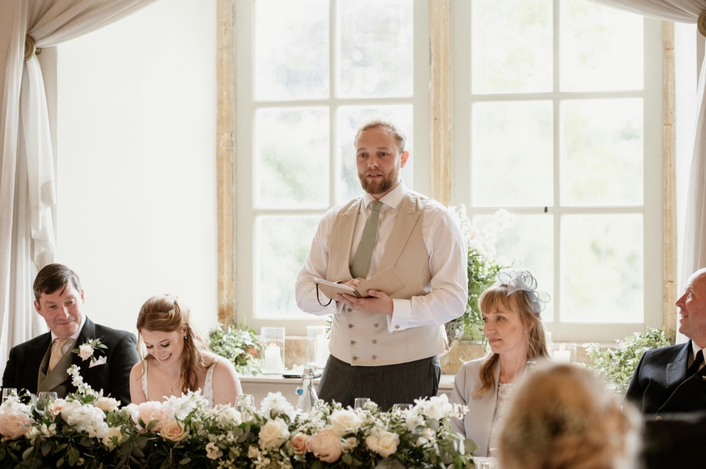 A groom giving a speech at a formal indoor wedding celebration with family and floral decorations