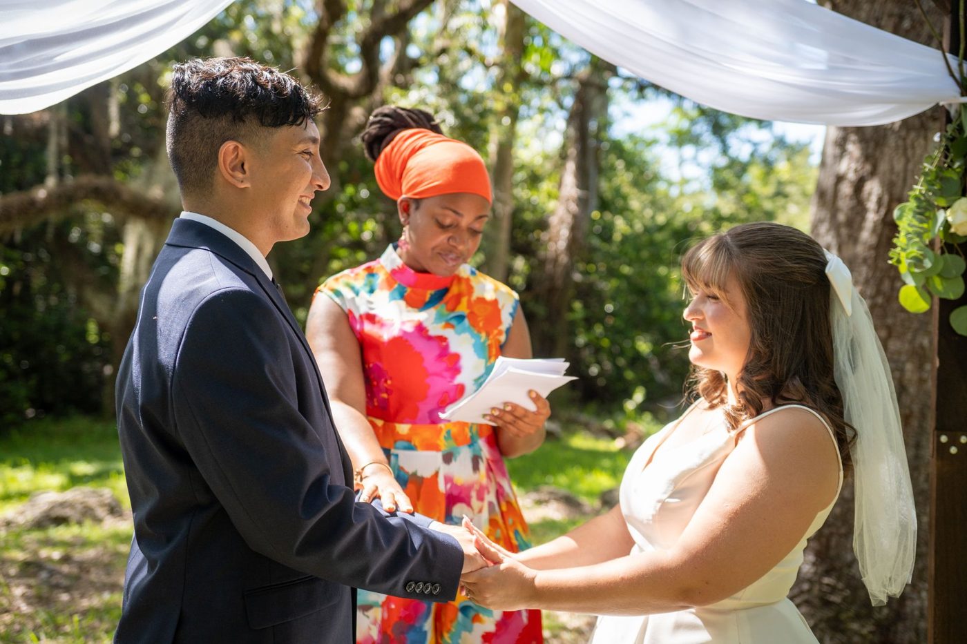 Couple exchanging rings and vows under trees while an officiant holds the ceremony script